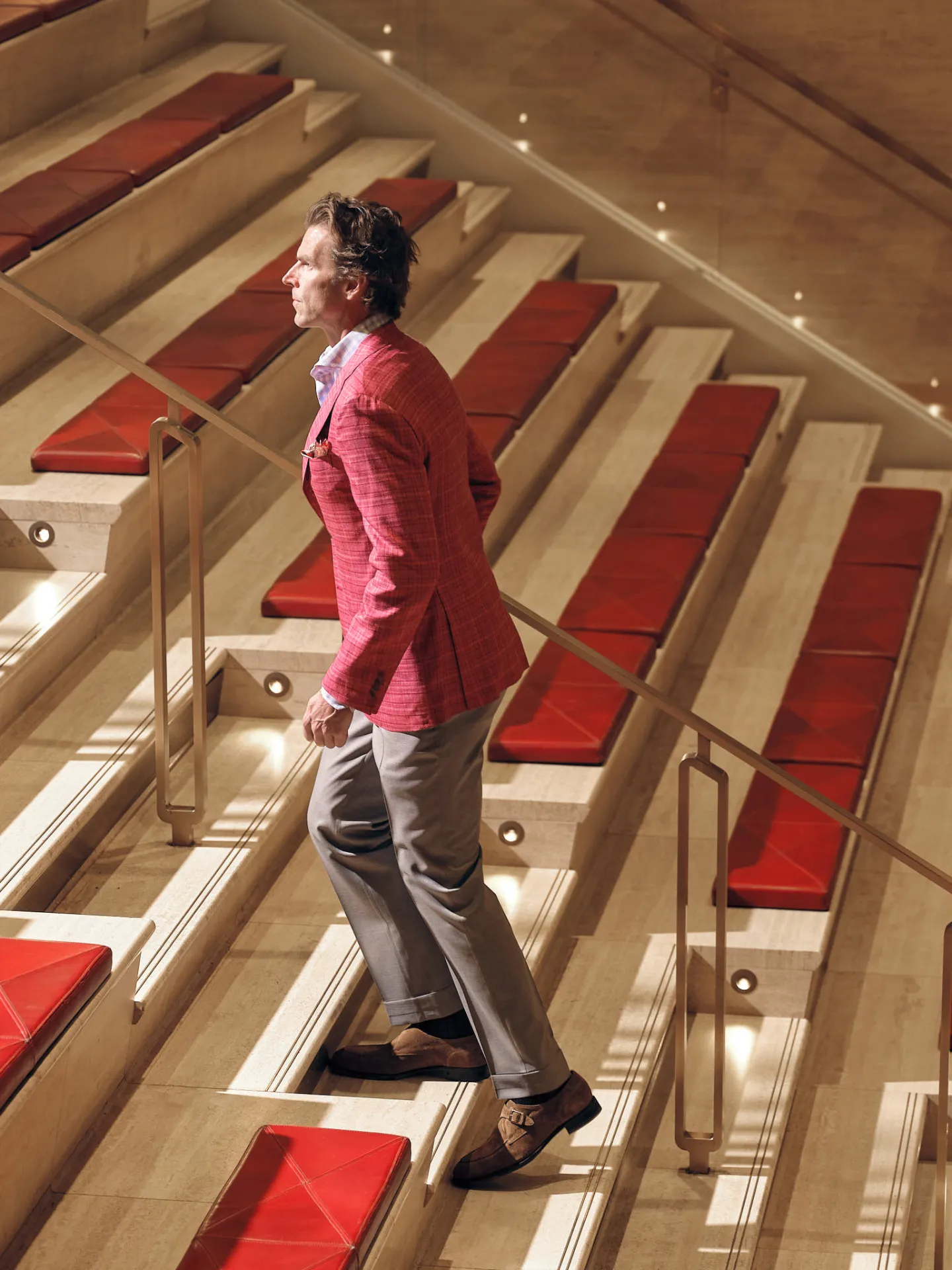 Man in a red checked sport coat and grey trousers walking up a stone staircase with red leather seats