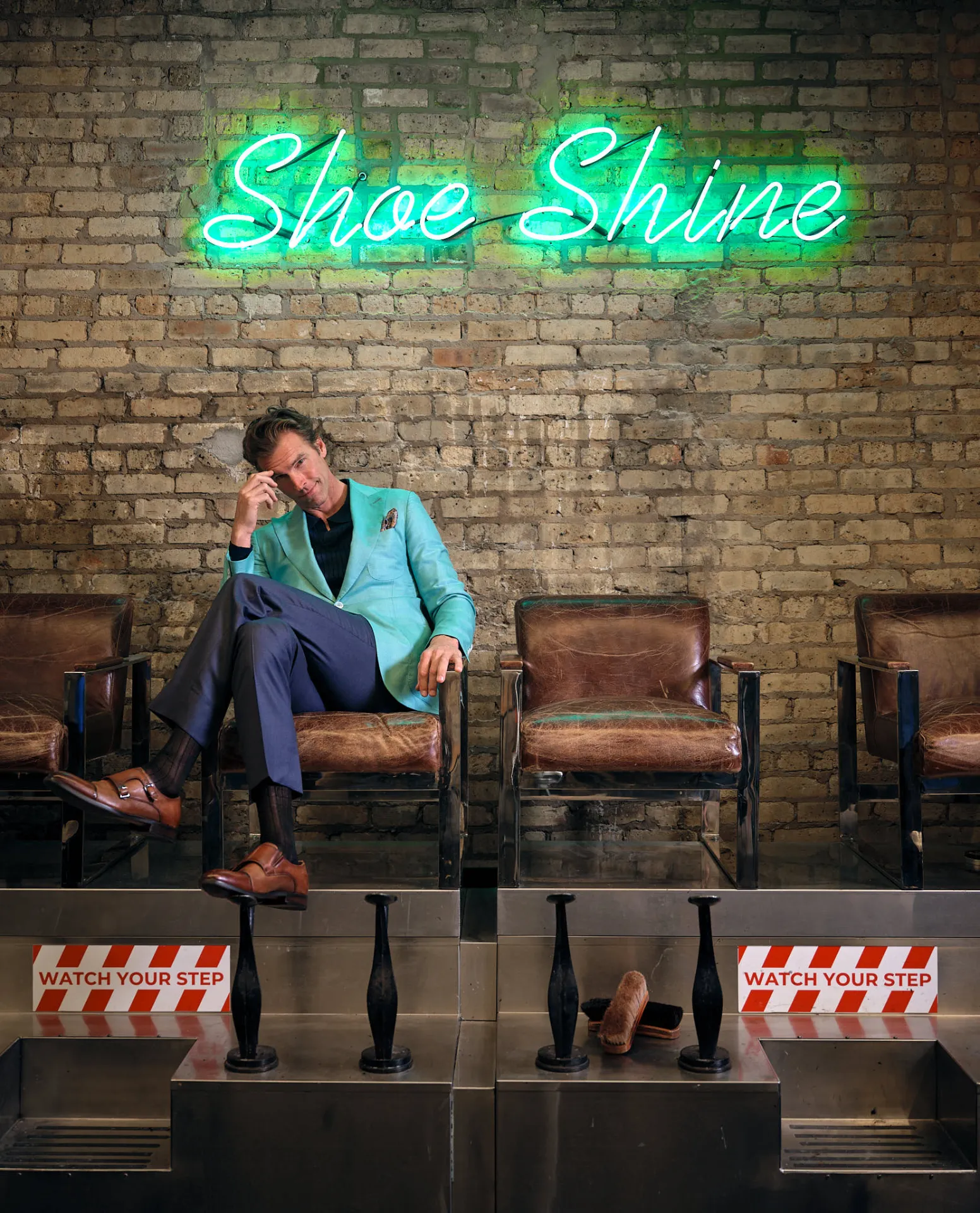 an in a mint green sport coat and navy trousers seated in a leather shoeshine chair under a green neon "Shoe Shine" sign