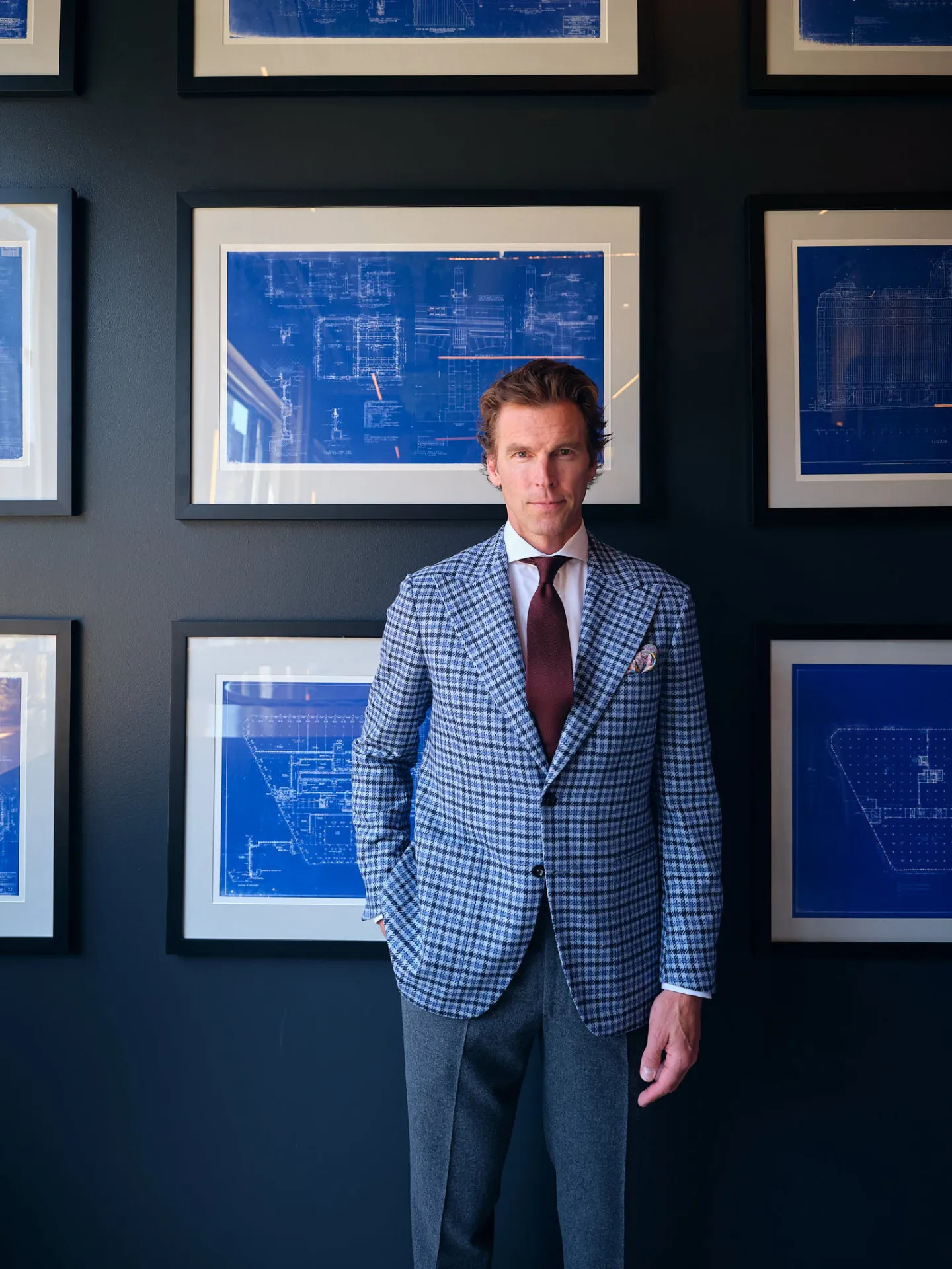 Man in a blue and navy houndstooth sport coat, white shirt and burgundy tie standing before framed blueprints
